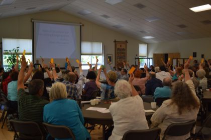 Congregation at tables in fellowship hall holding up orange voting slips for renewing our Welcoming Congregation status, from back 2017-5-21